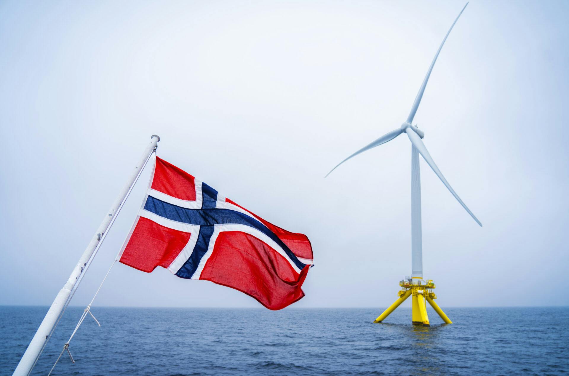 Norsk flagg blafrer med en havvindmølle i bakgrunnen.