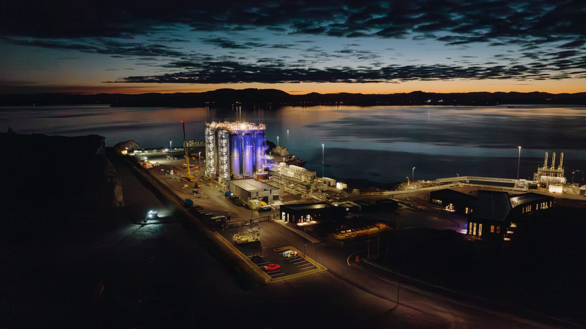 Industrianlegg ved en rolig vannkant i skumringen, med skarpe lys som lyser opp strukturer og overskyet himmel.