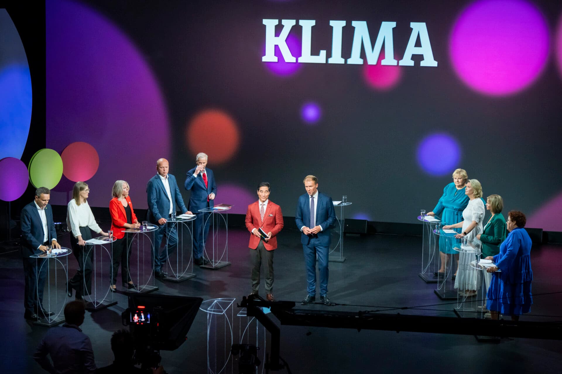 En gruppe mennesker står på podier på en scene under ordet «KLIMA» og deltar i en TV-sendt debatt eller diskusjon om klima.