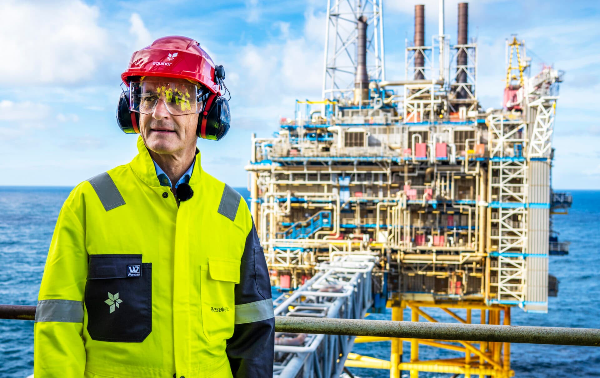 En person iført godt synlig verneutstyr og rød hjelm står på en oljeplattform til havs, med industristrukturer i bakgrunnen.