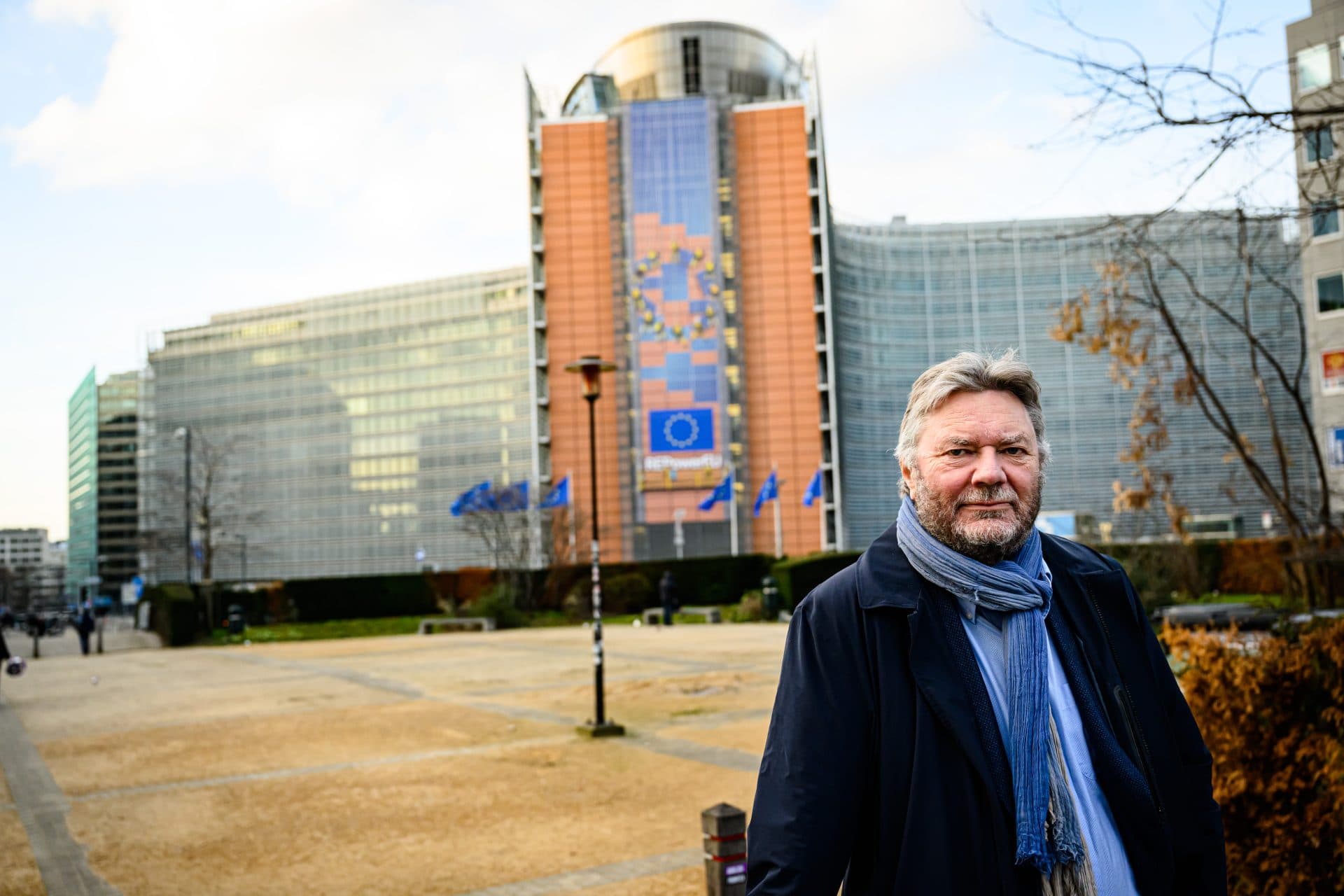 En mann i frakk og skjerf står foran en stor, moderne bygning med EU-flagg og et veggmaleri, sannsynligvis Europakommisjonens hovedkvarter.