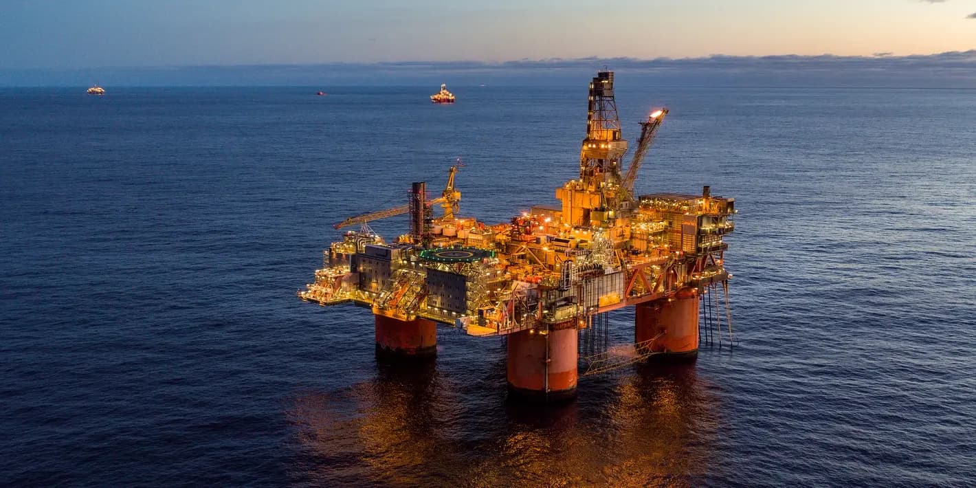 En opplyst oljeplattform ligger i havet i skumringen, med noen fjerne rigger synlige i horisonten under en delvis overskyet himmel.