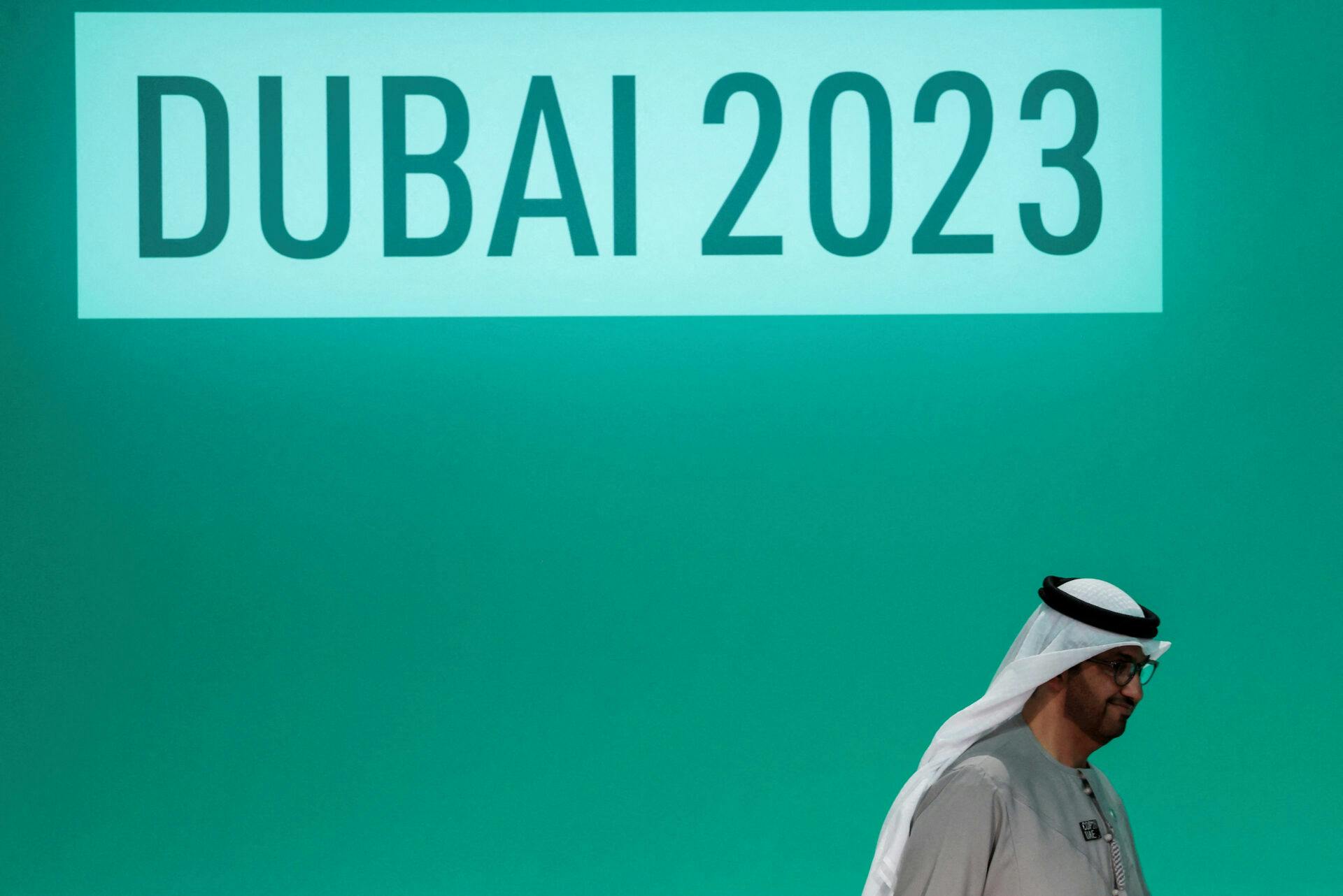 Mann i tradisjonelt saudiarabisk bekledning foran en stor grønn skjerm hvor det står Dubai 2023