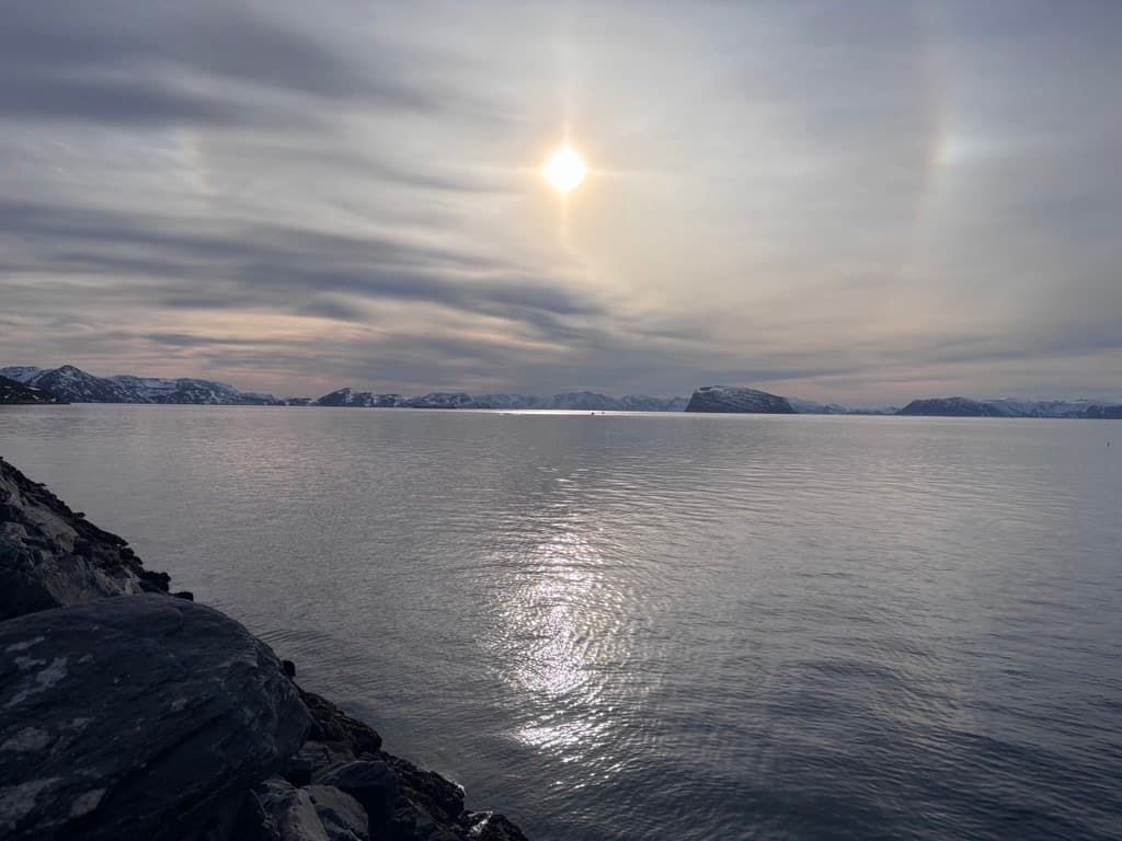 Solen skinner gjennom skyene over et rolig, vidstrakt vannspeil, med snødekte åser i det fjerne og en steinete kystlinje i forgrunnen.