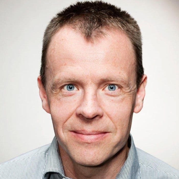 En middelaldrende mann med kort lysebrunt hår og blå øyne ser rett inn i kameraet, iført en lysstripet skjorte mot en ensfarget bakgrunn.