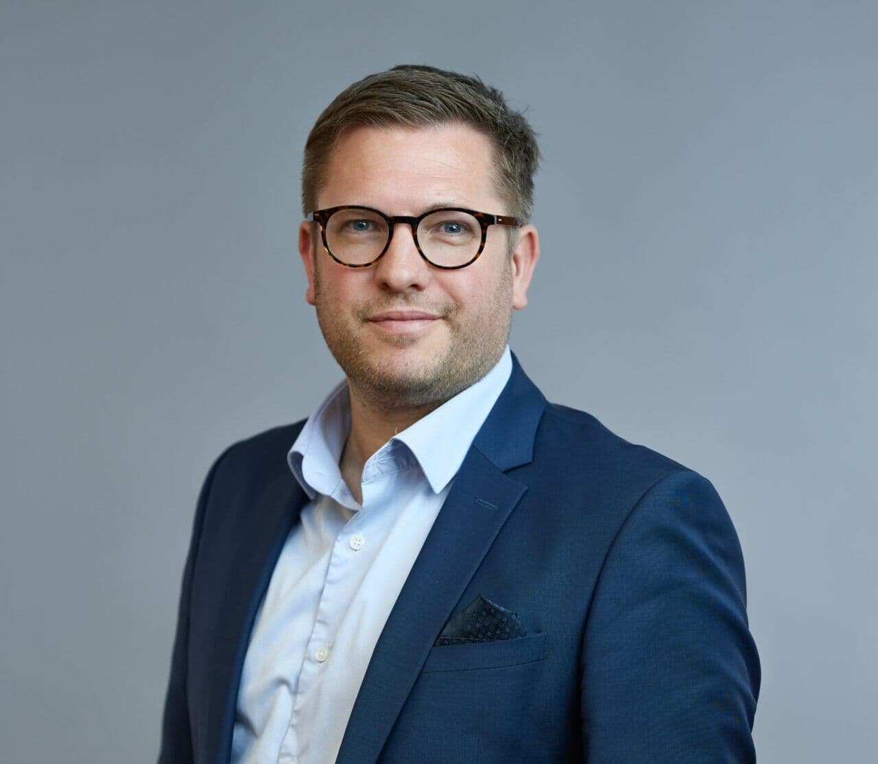 En profesjonell mann i blå dress og briller, som står mot en grå bakgrunn og smiler lett til kameraet.