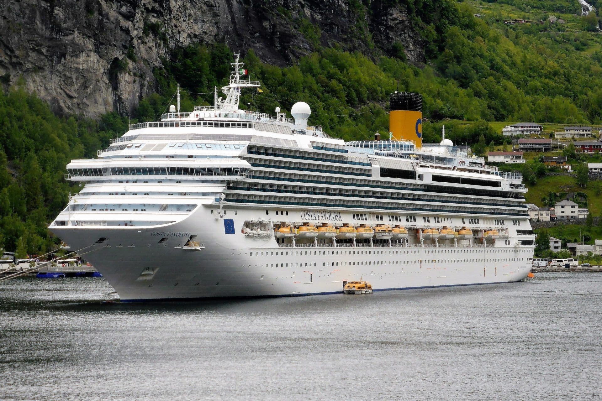 Et stort, hvitt cruiseskip ligger ved kai i en rolig havn nær en grønn, fjellrik kystlinje med spredte hus.