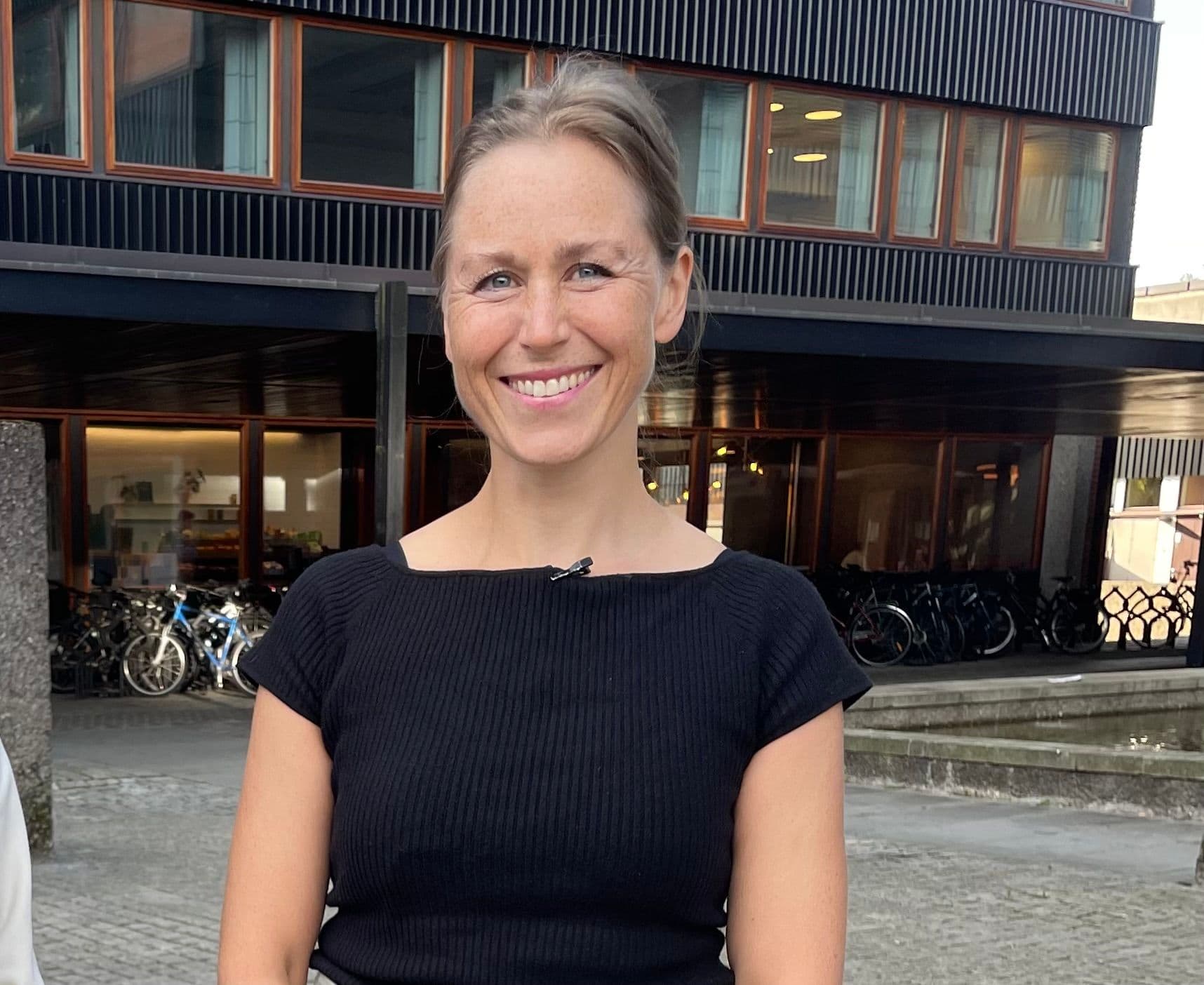 En kvinne med bakoverbøyd, lysebrunt hår og svart topp står utendørs foran en moderne bygning med sykler parkert i nærheten.