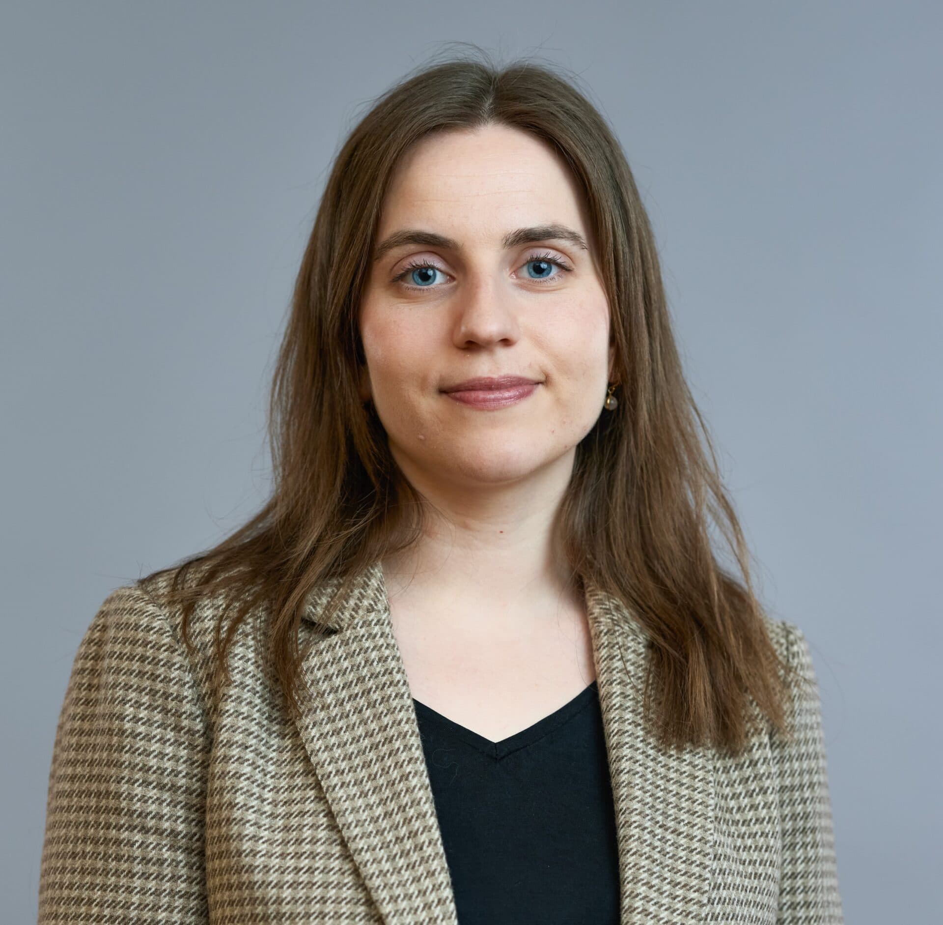 Profesjonelt portrett av en kvinne med middels langt brunt hår, iført en lys dressjakke og svart topp, mot en grå bakgrunn.