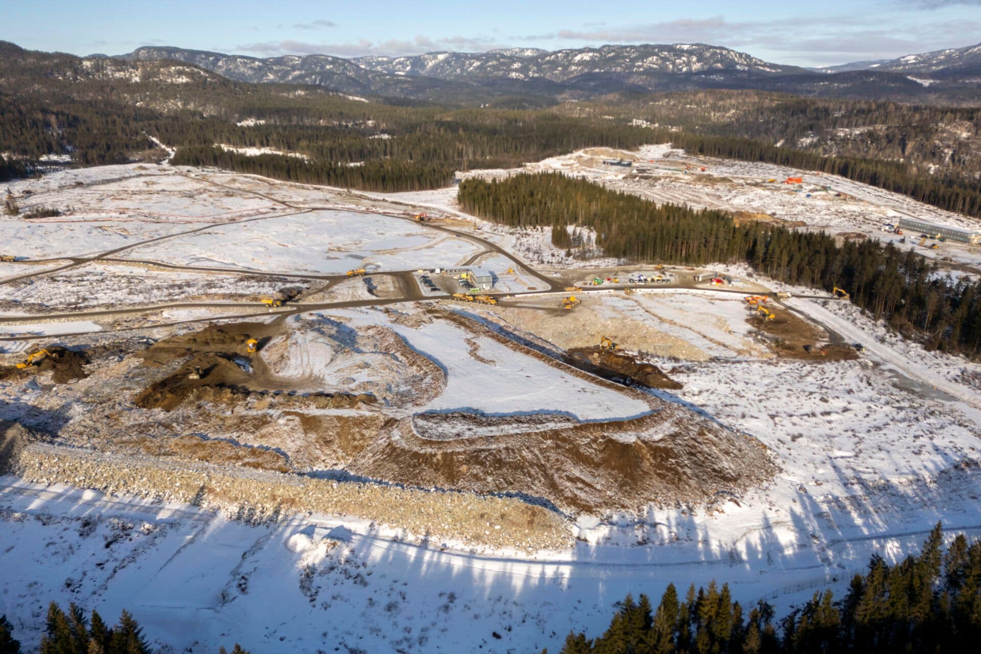 En snødekt byggeplass med maskiner og utstyr ligger i et skogkledd landskap, omgitt av fjell i bakgrunnen.