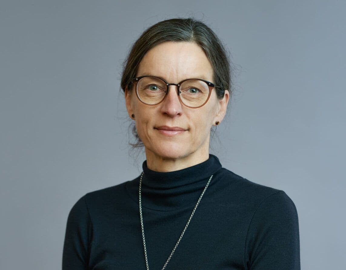 A woman with glasses and a ponytail, wearing a black turtleneck and a necklace, stands against a grey background and looks directly at the camera.